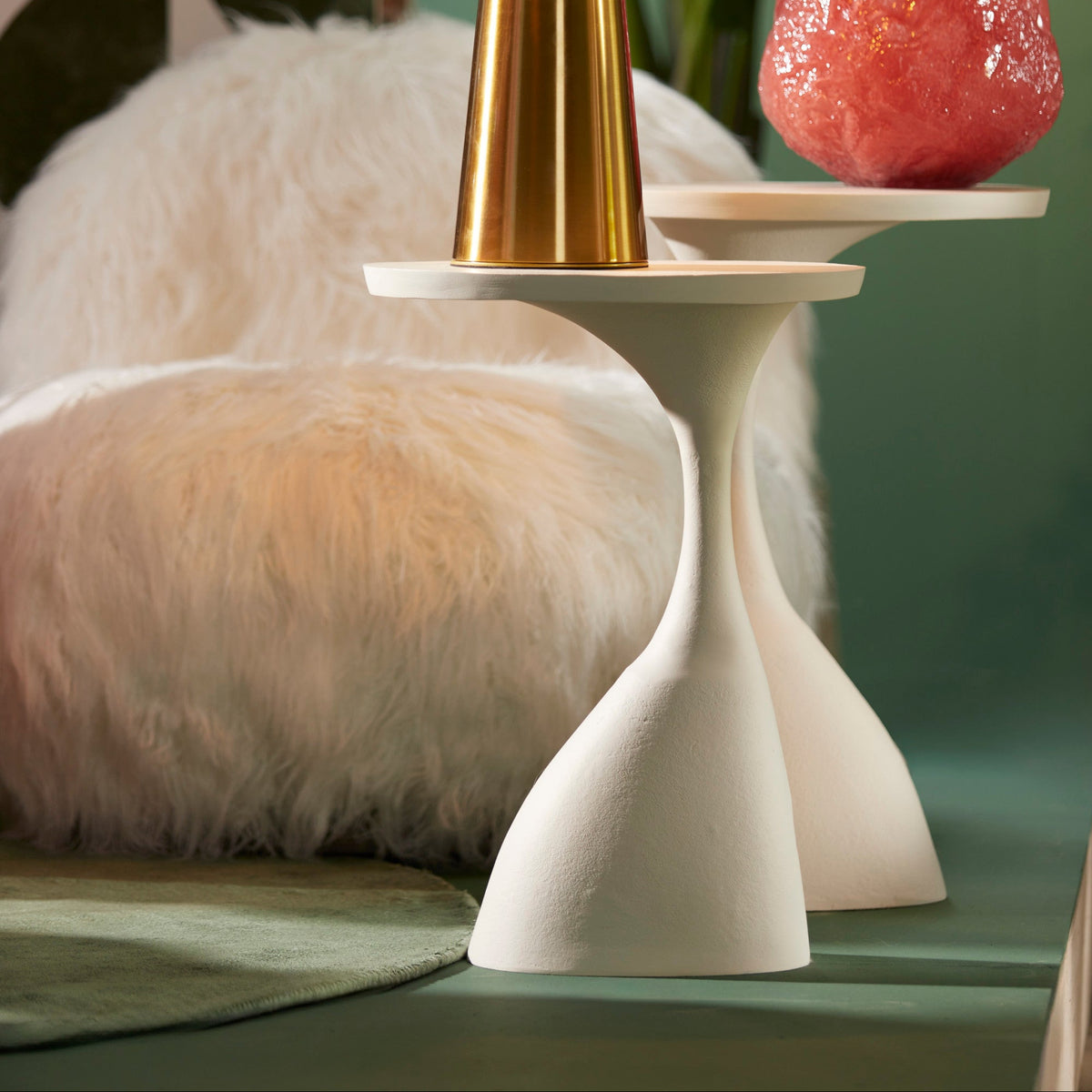 Gold table lamp on a white pedestal with a pink vase and plant in the background.