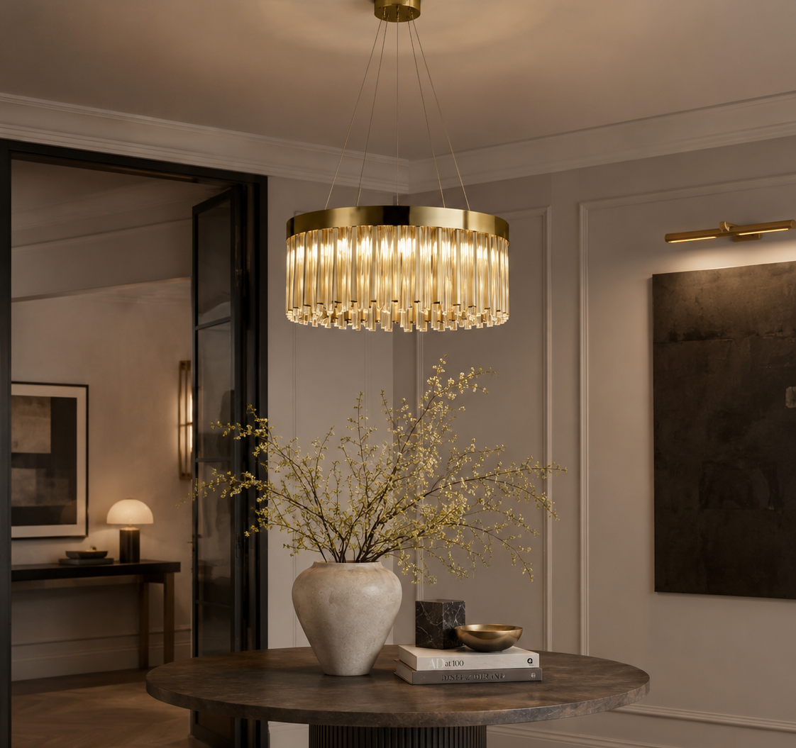 A modern cylindrical pendant chandelier with a gold-toned frame and clear glass or crystal vertical bars, hanging over a round dark wood dining table in a styled interior room.