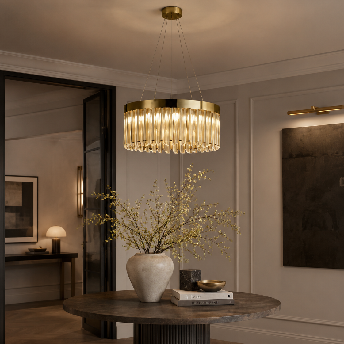 A modern cylindrical pendant chandelier with a gold-toned frame and clear glass or crystal vertical bars, hanging over a round dark wood dining table in a styled interior room.