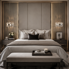 A luxury bedroom featuring a large bed with neutral gray and white bedding, a matching upholstered bench at the foot, a tall padded headboard, and symmetrical crystal chandeliers on dark wood nightstands.