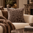 A styled living room scene featuring a textured brown throw pillow on a cream bouclé armchair, with a faux fur throw blanket draped over the seat. In the foreground, a marble coffee table holds books, a bowl, and a lit candle, with a fireplace and plants in the background.