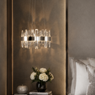 A modern multi-light chandelier with clear crystal droplets and silver-toned metal accents, hanging in a luxury bedroom above a dark wood nightstand with a vase of white flowers.