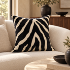A square throw pillow with a black and cream zebra stripe pattern, resting on a textured beige sofa in a styled living room setting.